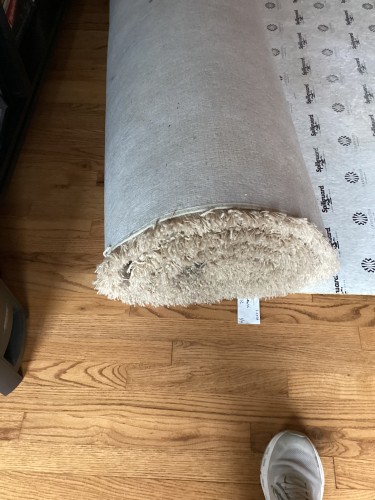Close-up of a rolled residential rug, showing its underside and a SpillGuard rug pad on a wooden floor, indicating preparation for professional rug cleaning service.