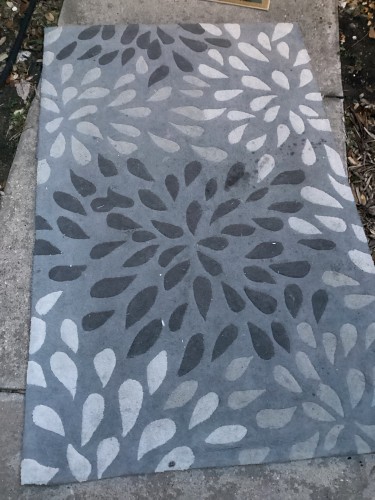 A large grey residential area rug with a faded floral or leaf pattern, placed on a concrete surface, indicating the need for or completion of professional rug cleaning and treatment for faded areas and potentially fringes.