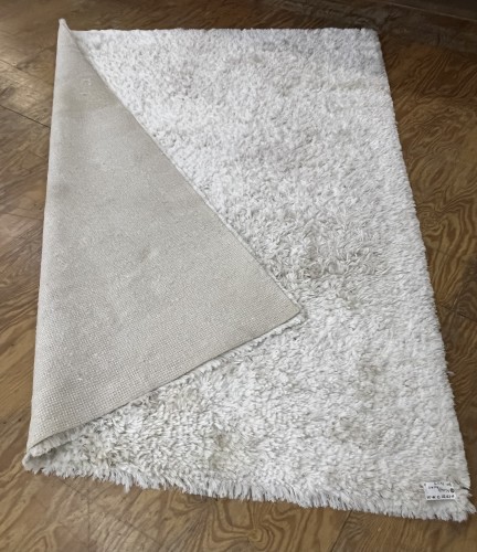 A clean white shag area rug, with one corner folded to show the beige non-slip backing, resting on a wooden floor after professional residential cleaning by Dalworth Rug Cleaning.