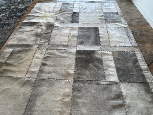 A large residential patchwork cowhide rug with various shades of grey and beige showing visible heavy to moderate soil and faded spots on a wooden floor before professional cleaning.