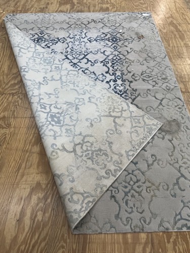 Residential area rug from Dalworth Rug Cleaning, a light-colored patterned rug on a wooden floor, with a section folded over, highlighting areas of discoloration or potential staining and damage in a corner, relevant to the cleaning service where 100% stain removal was not guaranteed.