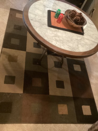 A geometric square-patterned area rug in shades of brown, beige, and olive green, completely deep-cleaned and pile-restored to eliminate furniture marks beneath a round coffee table.
